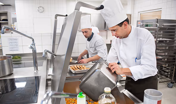 Zwei Köche mit Kochmützen bei ihrer Arbeit in der Großküche eines integrativen Betriebs.