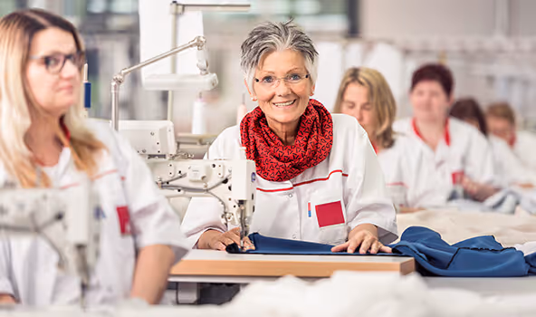 Eine Näherin, angestellt in einem integrativen Betrieb, sieht von Ihrer Arbeit auf und lächelt in die Kamera.