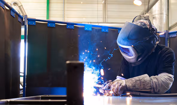 Ein Arbeiter eines integrativen Betriebs mit Schutzkleidung und Schweißerbrille schweißt Metall in einem abgetrenntem Bereich einer Fertigungshalle.