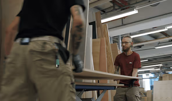 Tobias, Tischler in einem integrativen Betrieb, wie er mit einem Arbeitskollegen ein großes Holzbrett transportiert.