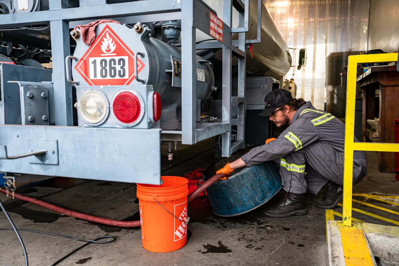 Worker draining liquid from hazardous materials tanker labeled “1863” into orange bucket, wearing gloves and reflective coveralls.