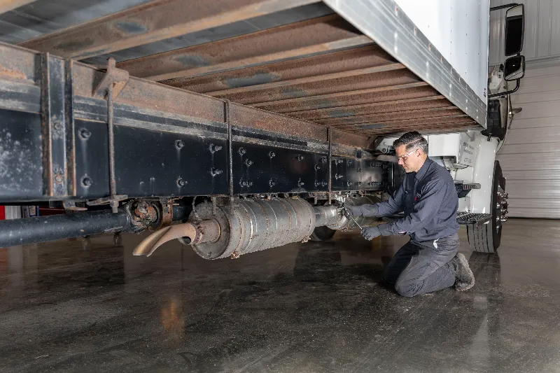 Technician inspecting the diesel aftertreatment system underneath a heavy-duty truck during preventive maintenance to improve emissions performance and reliability.