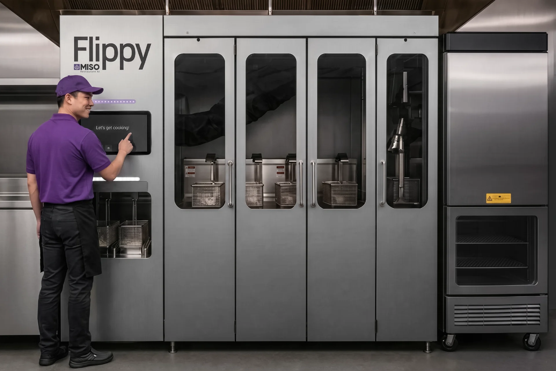 A worker in a purple shirt operates the touchscreen of a large, stainless steel Flippy robotic frying system.