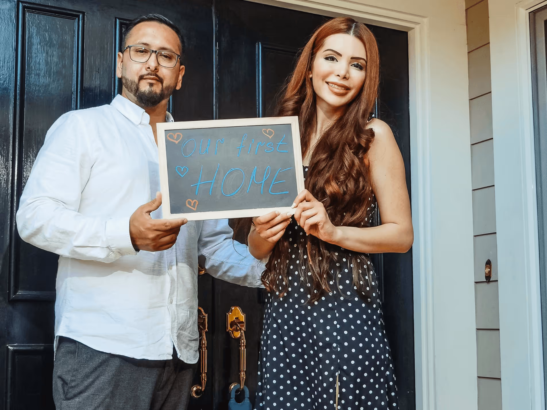 A couple holding a board that says "our first home" in front of the door