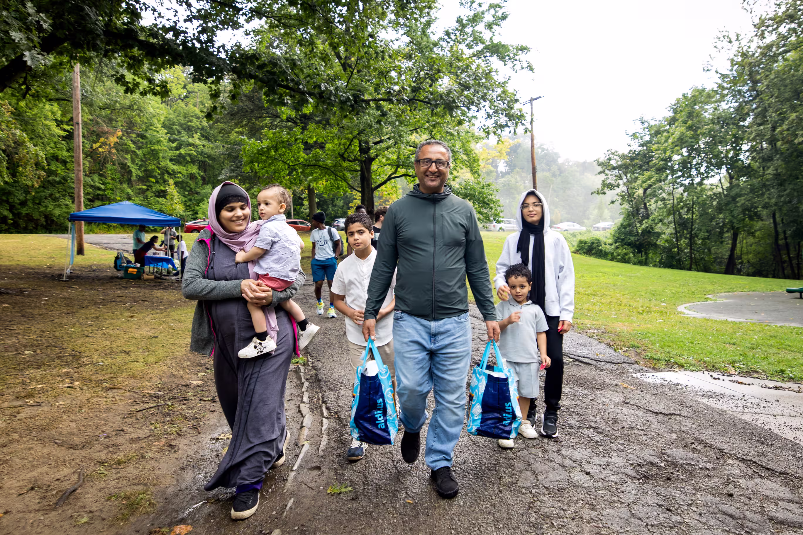 A large smiling family walking down a damp asphalt path surrounded by lush green trees.