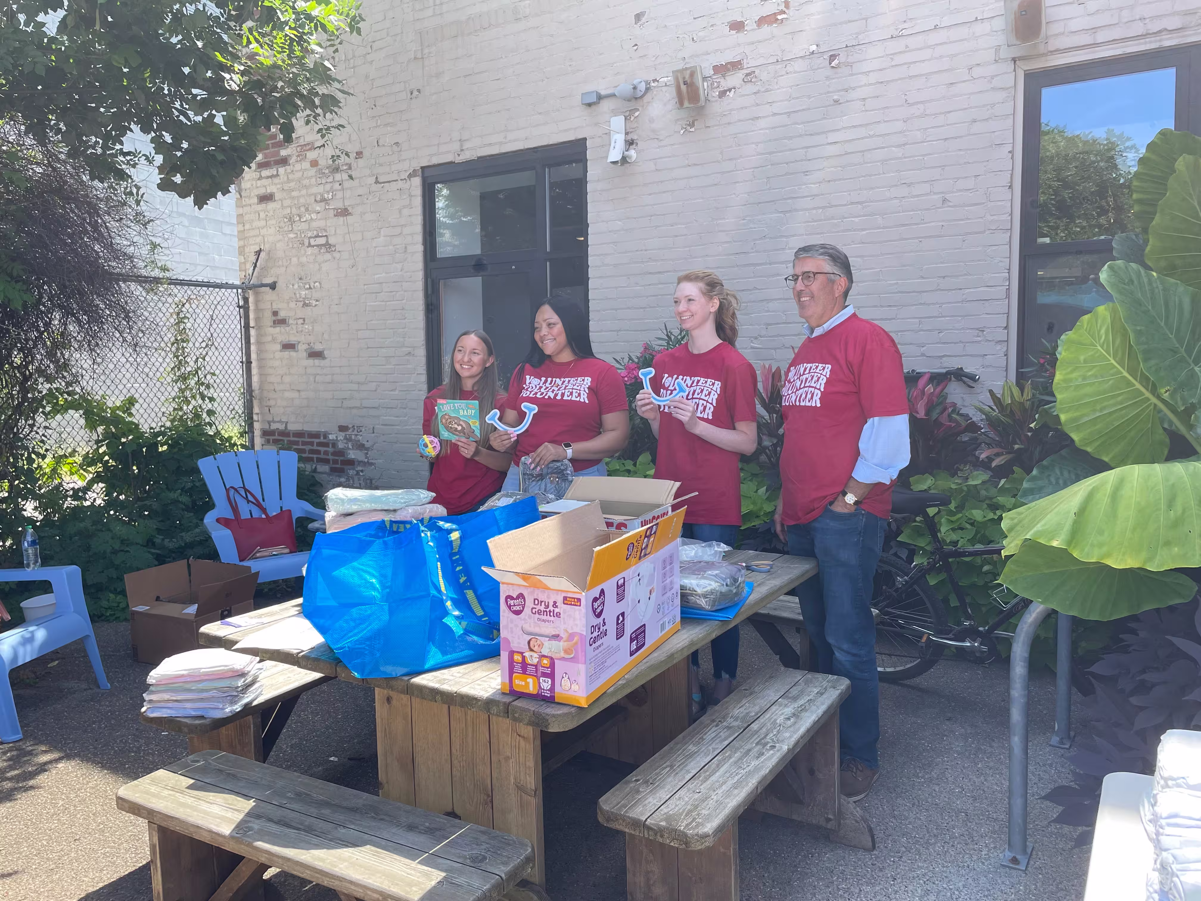 A group of four adults wearing matching red volunteer shirts smile for a photo holding various In-Kind donation items.