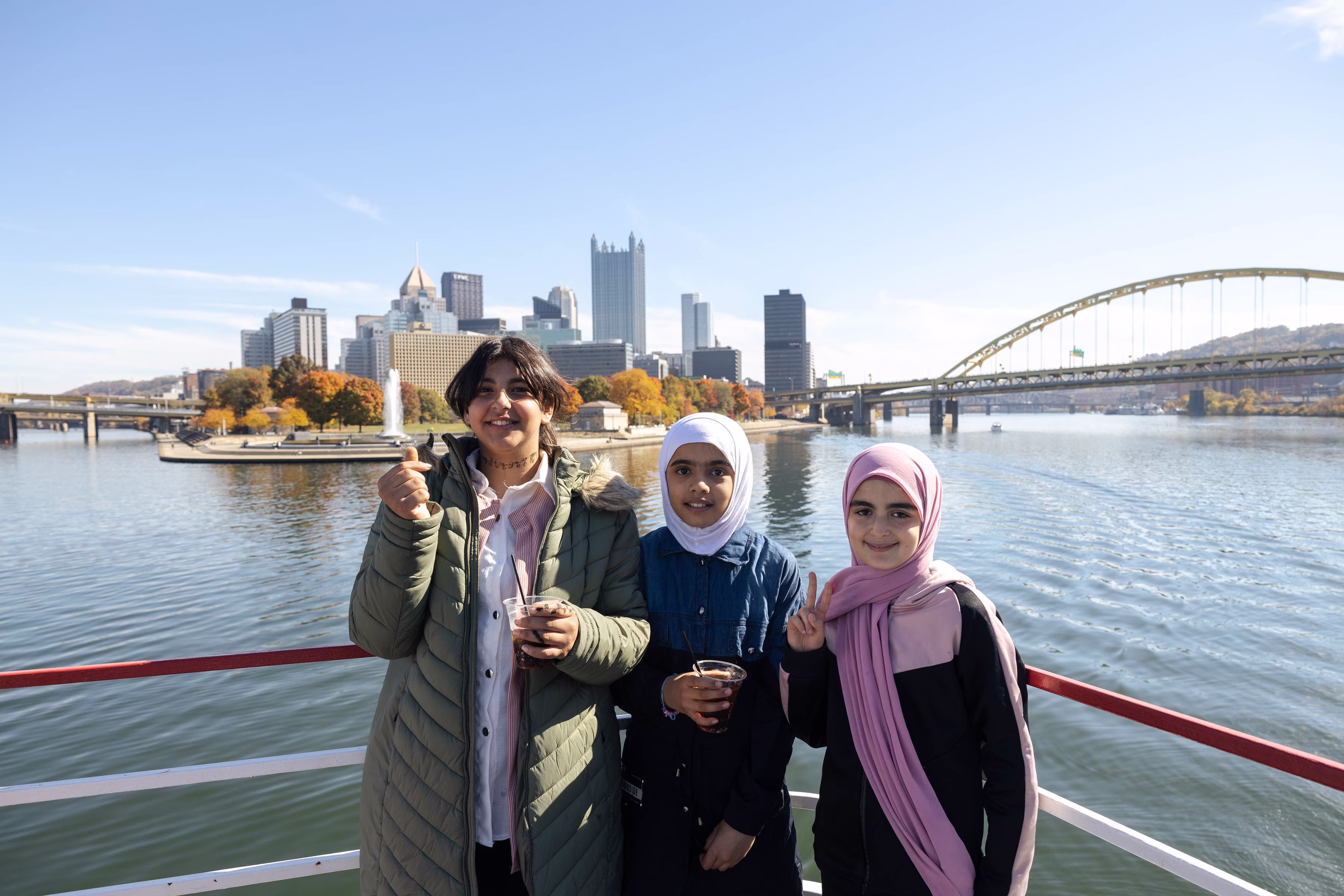 Three girls on the Gateway Clipper smile at the camera holding drinks, on a sunny day. Behind them is a view of Pittsburgh's Point State Park.