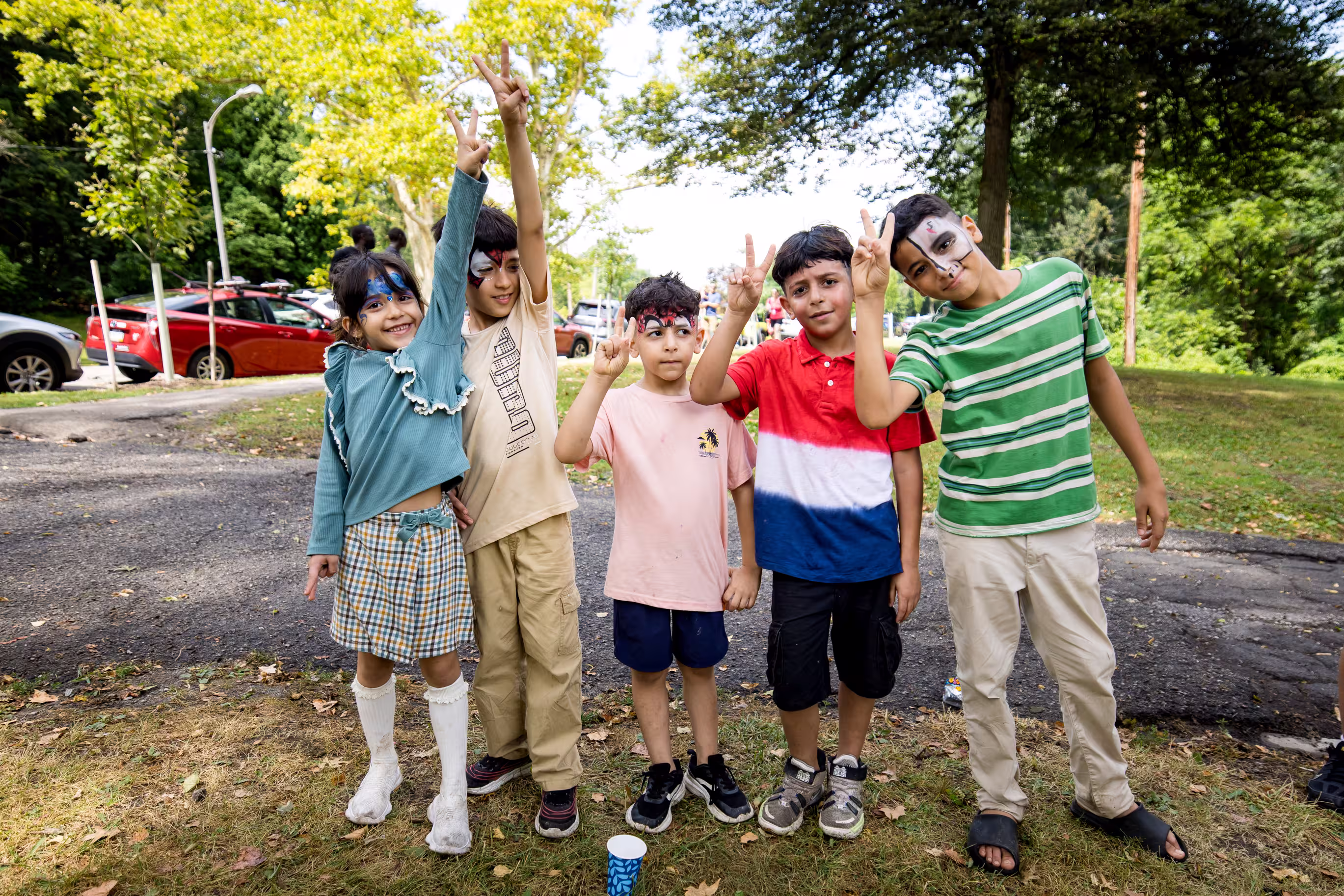 Five kids stand pictured in a park, smiling and holding up peace signs on a bright sunny day. Each of the kids have their face painted in a unique way.