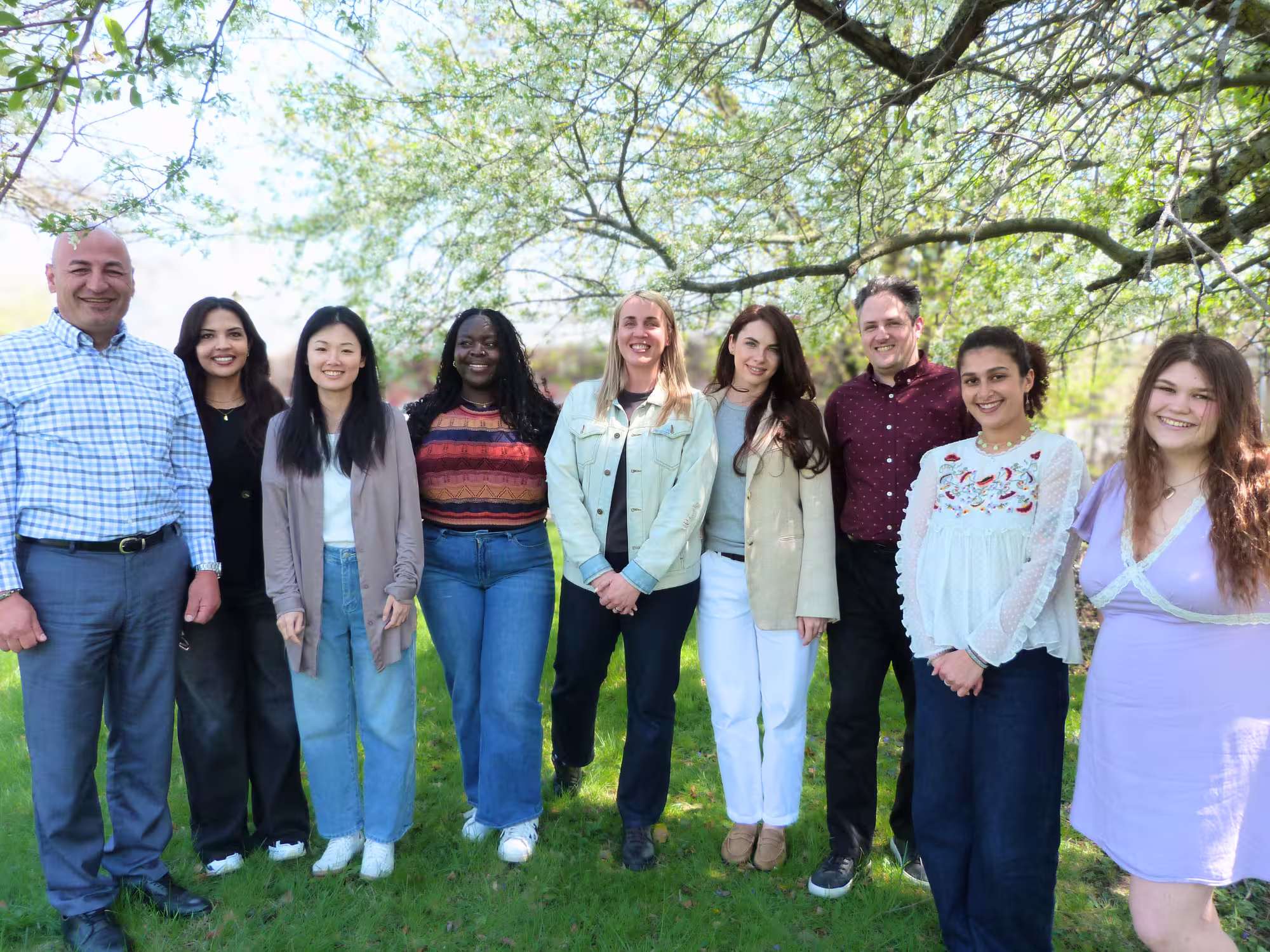 The Hello Neighbor Employment Services Team is pictured outside on a sunny day, standing together in a line, smiling, beneath a big leafy green tree.