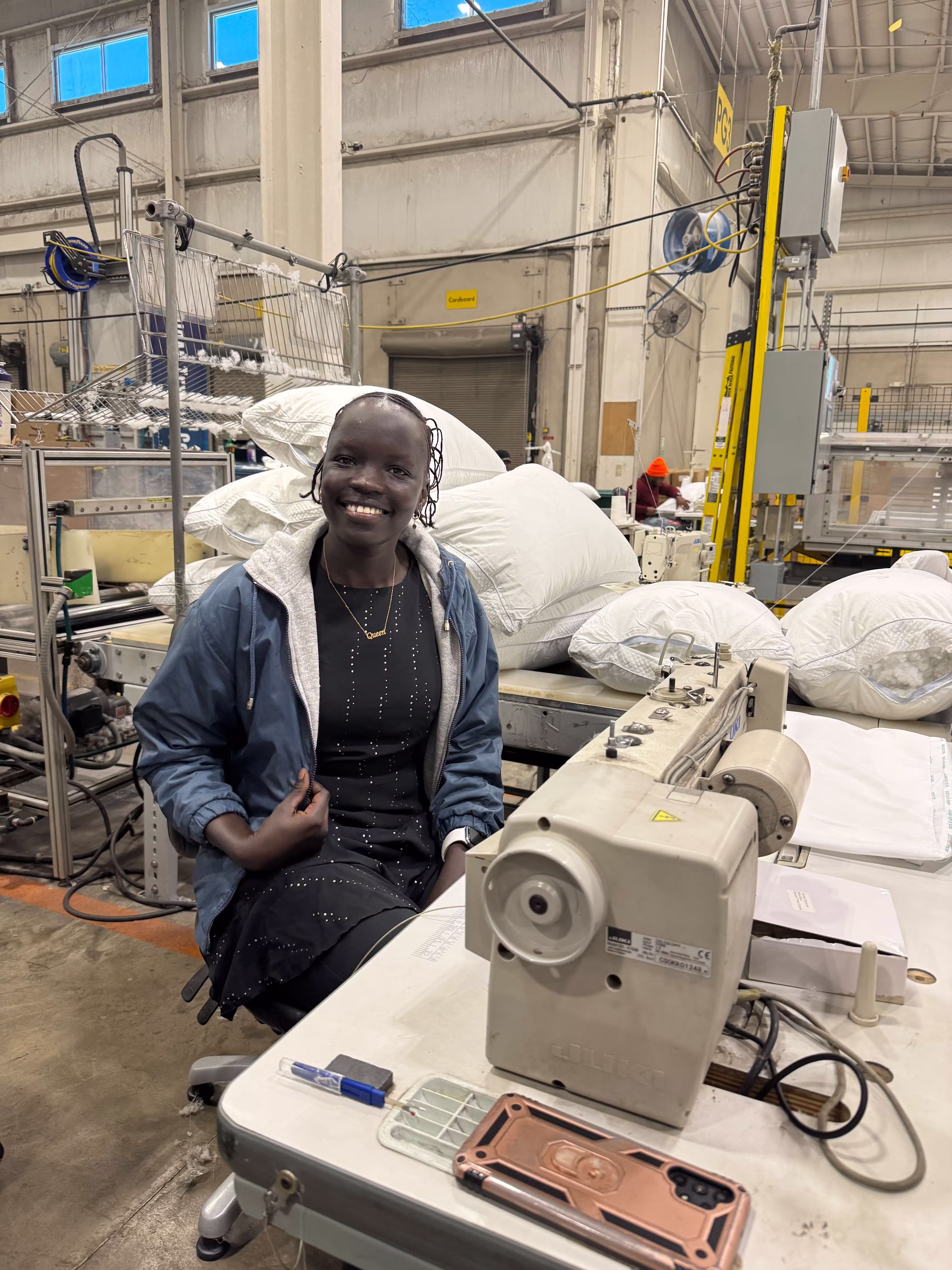 A smiling woman sits beside her sewing machine inside a textile manufacturing space.