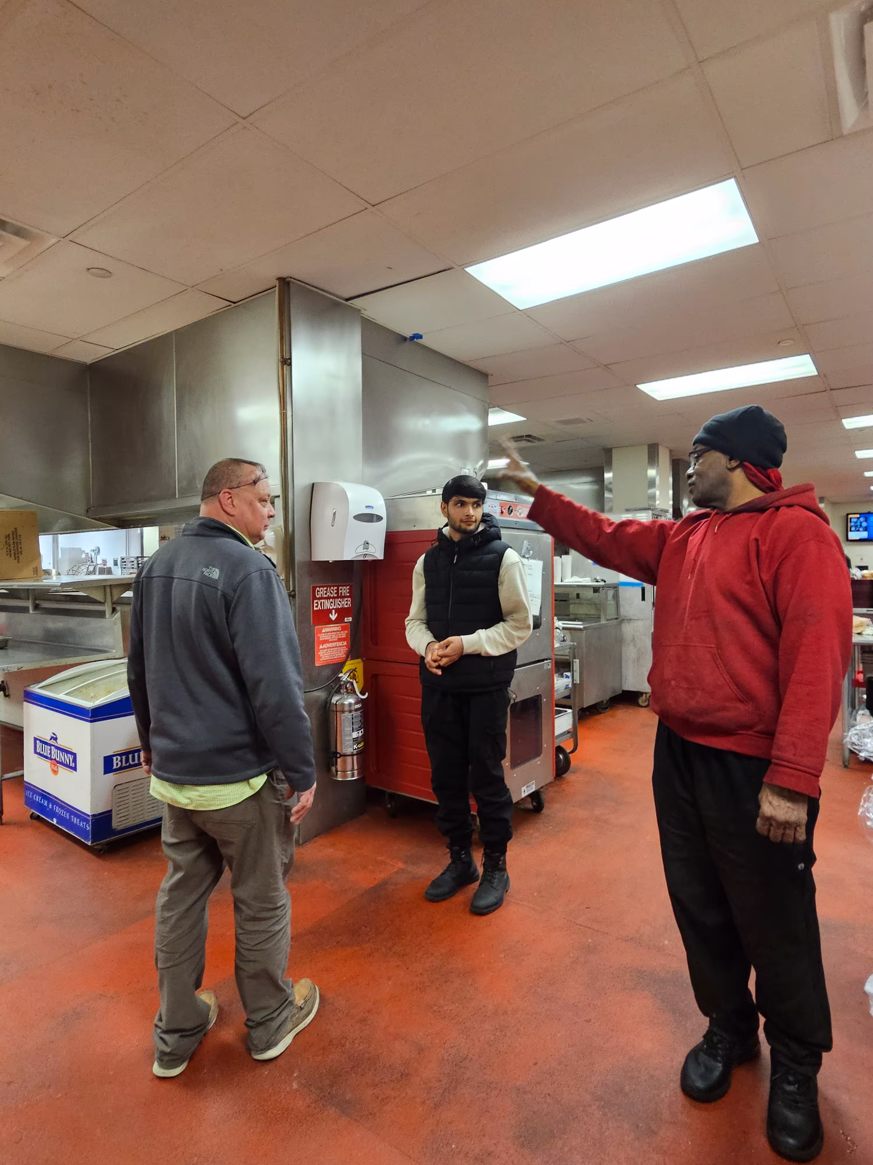 Three men stand in a kitchen, listening to instruction from another employee.