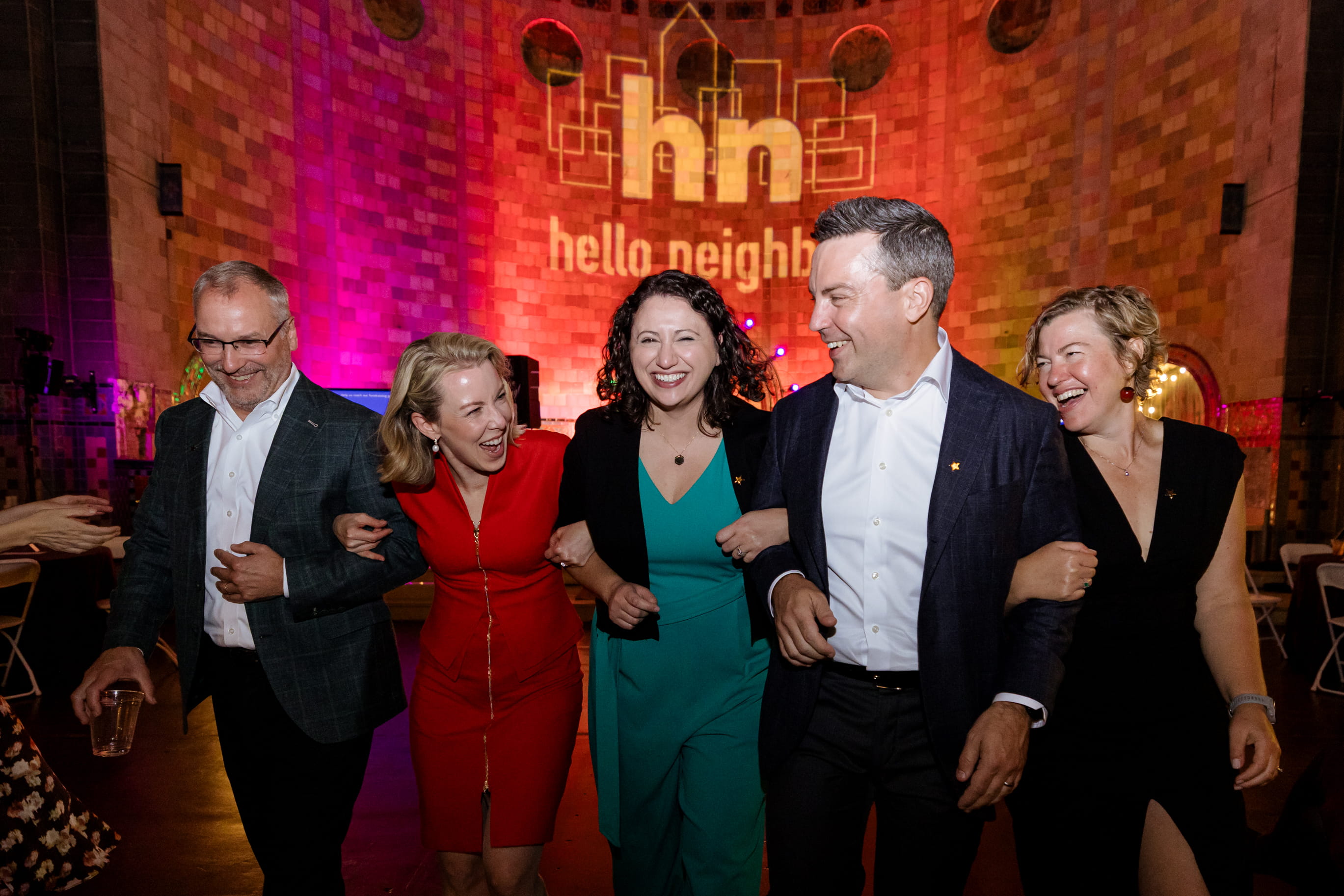 Attendees at The Neighborhood Nosh laughing and line dancing in front of illuminate brick wall emblazoned with "Hello Neighbor"
