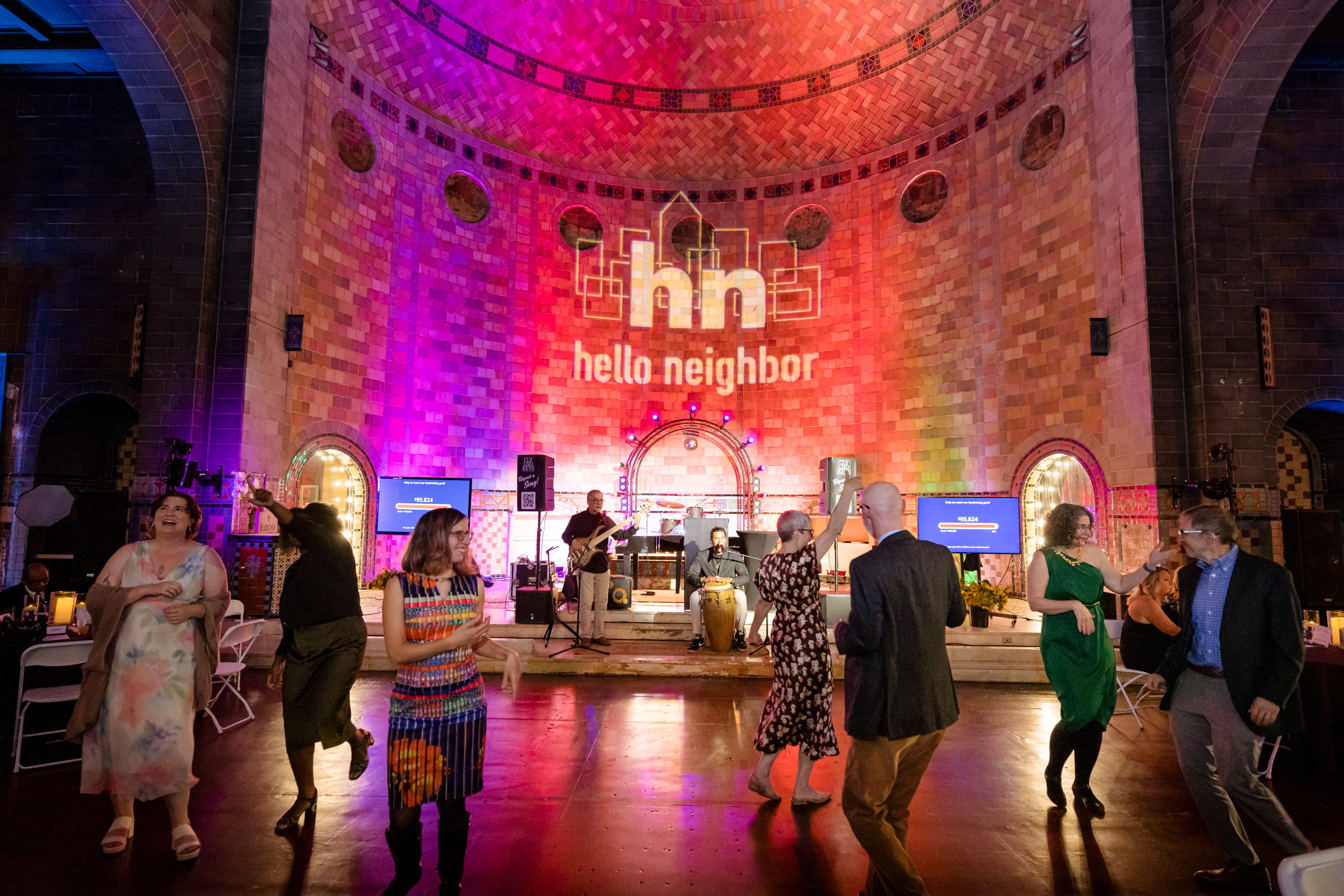 Attendees dancing at The Neighborhood Nosh in front of stage, band, and illuminated Hello Neighbor logo on brick wall