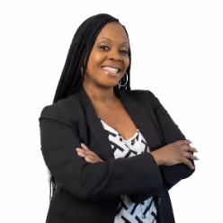 Smiling woman with long braided hair wearing a black blazer and patterned blouse standing with arms crossed.