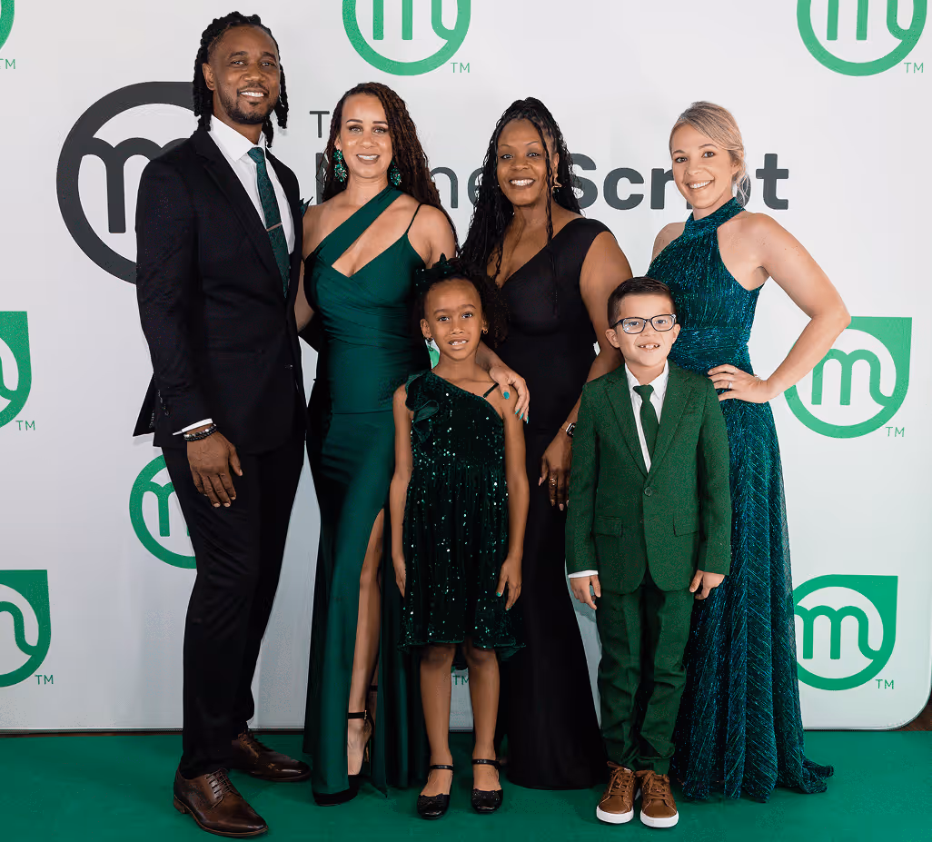 Five people dressed formally posing together, three women in black and green dresses, one woman in a pink dress, and one man in a black suit with a green tie.
