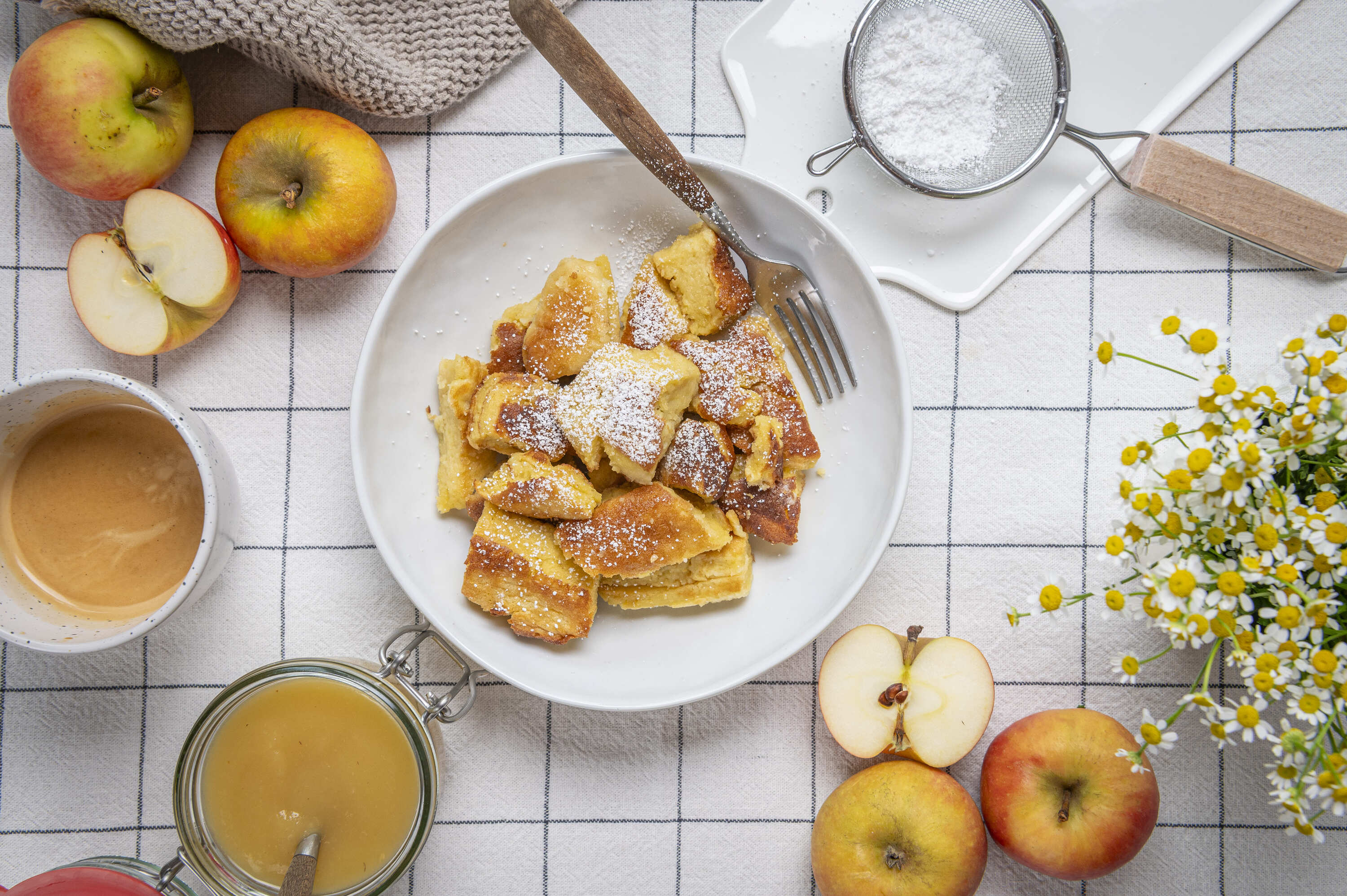 Auf einem Geschirrtuch steht eine Schüssel mit leckerem Kaiserschmarrn. Rundherum liegen Äpfel und ein Glas Apfelmus.