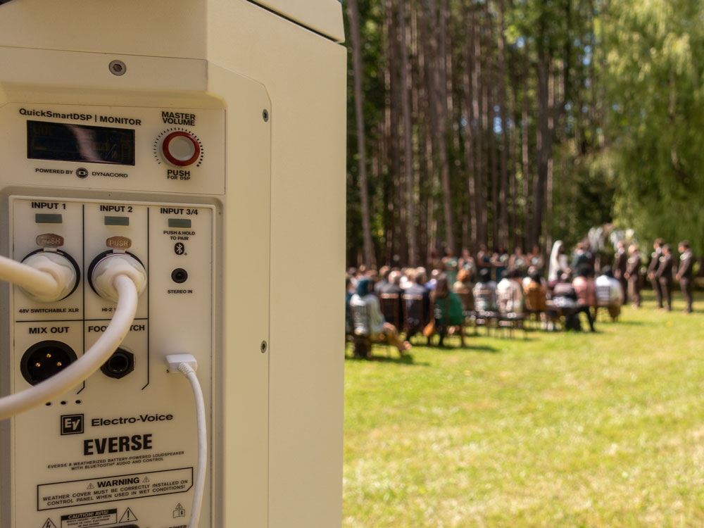 Pro audio setup — rear of a speaker in focus with an outdoor forest ceremony blurred behind; clear, reliable ceremony sound.