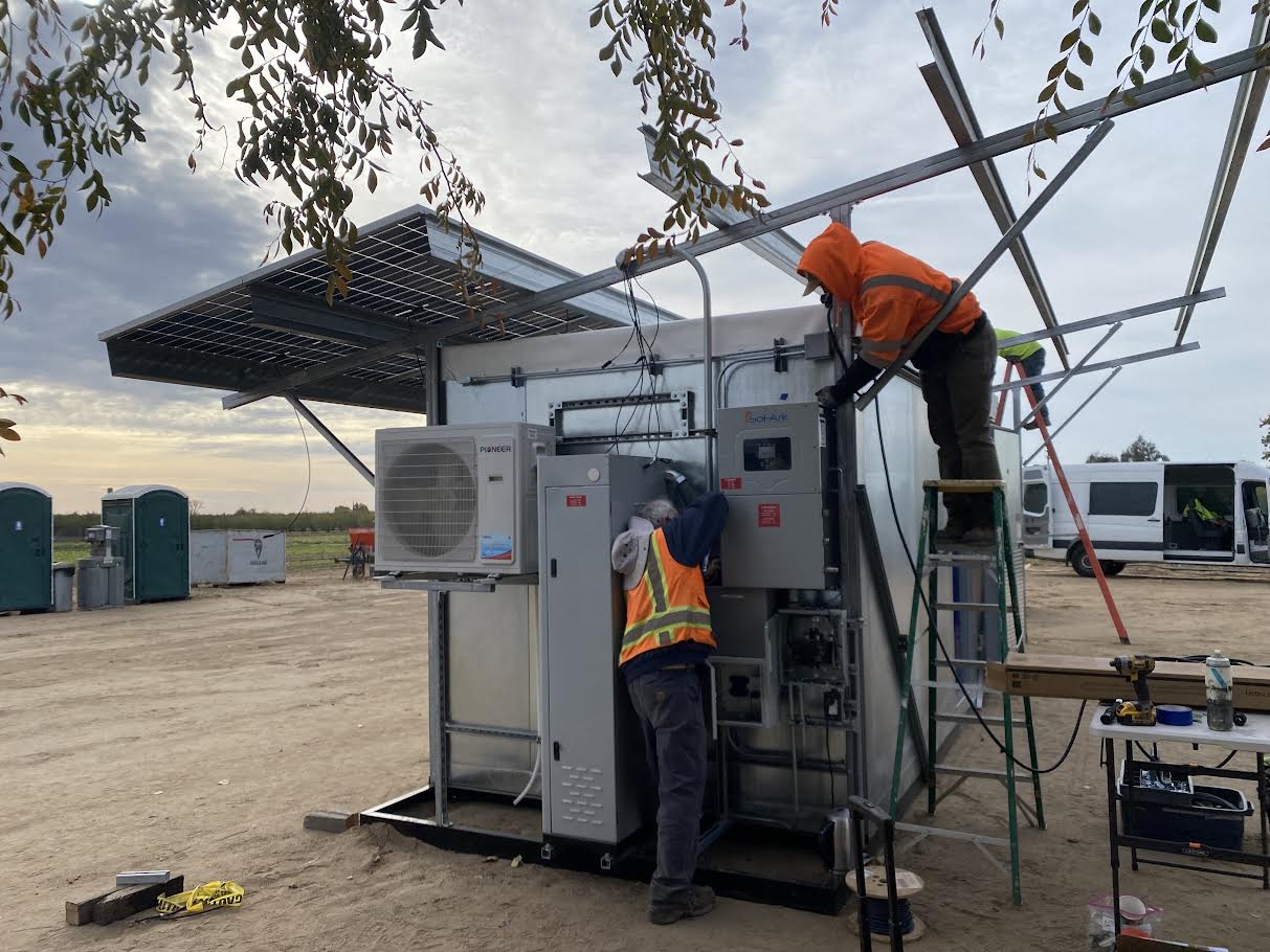 Two workers set up a solarfresco solar powered cooler in an unpaved part of a field 