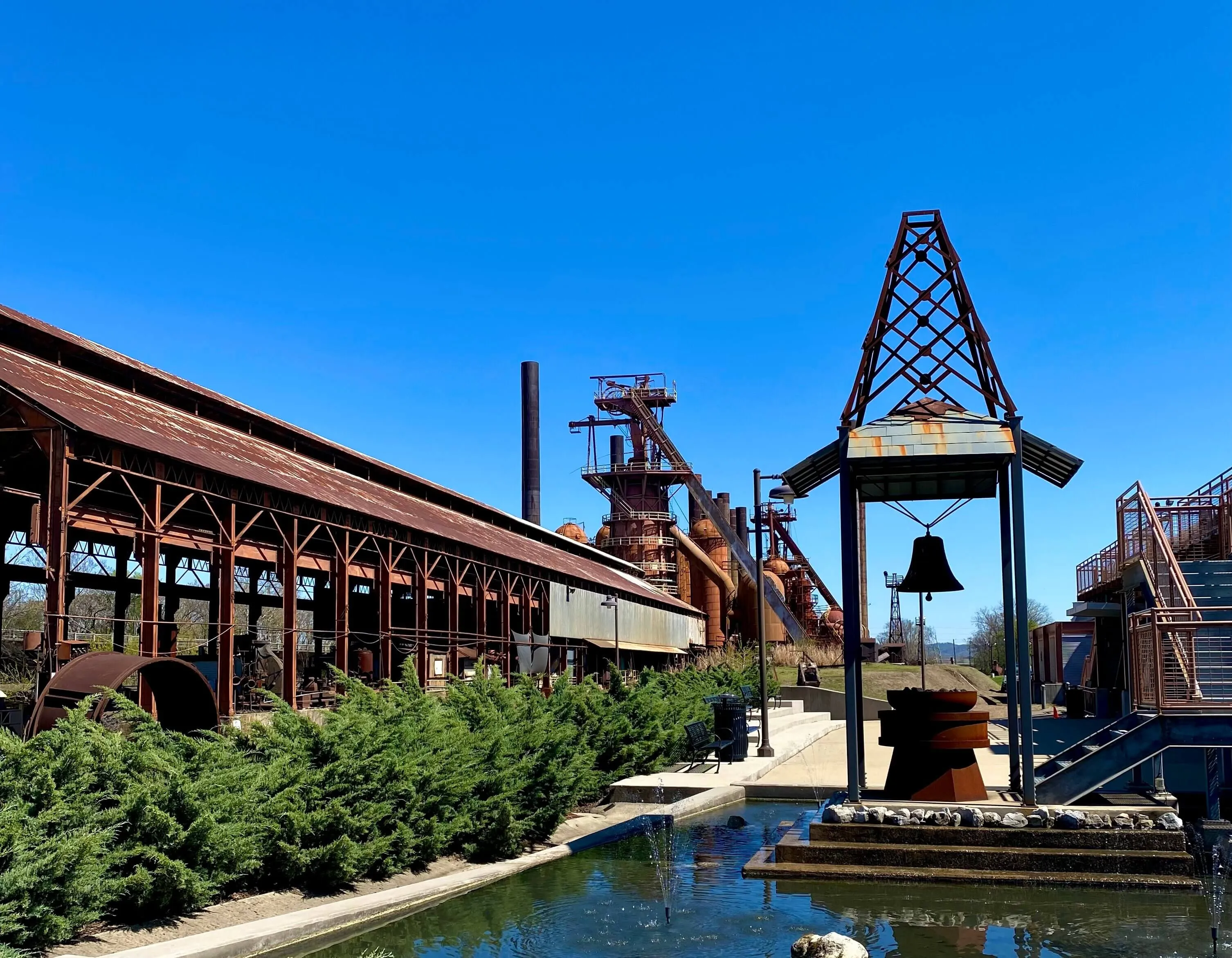 Outdoor scene featuring a rusty metal bell suspended under a metal frame near a rectangular water fountain with small jets, green shrubs alongside a long industrial building, and old factory structures with clear blue sky background.