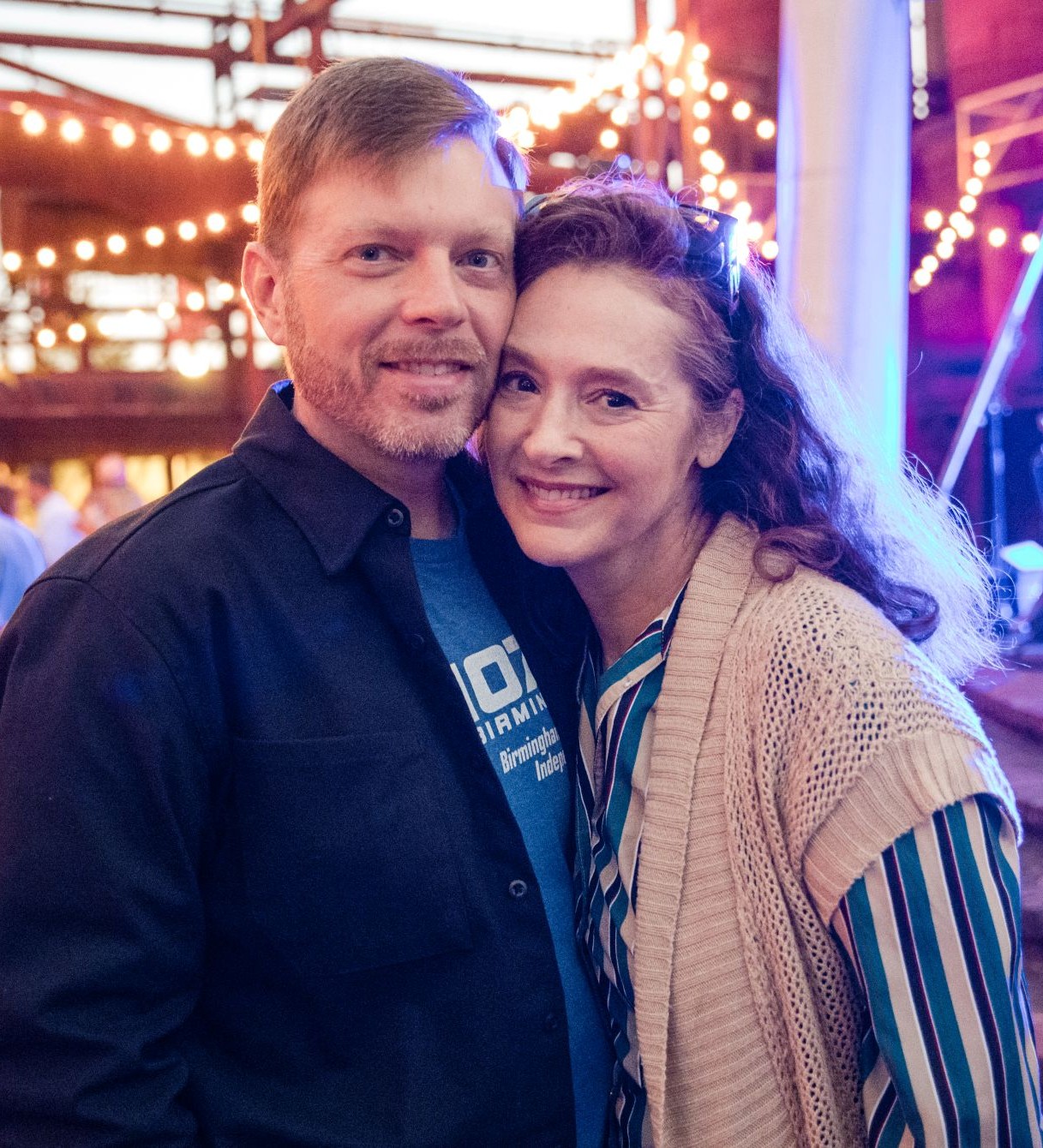 Smiling man and woman closely posing together in a warmly lit indoor setting with string lights in the background.