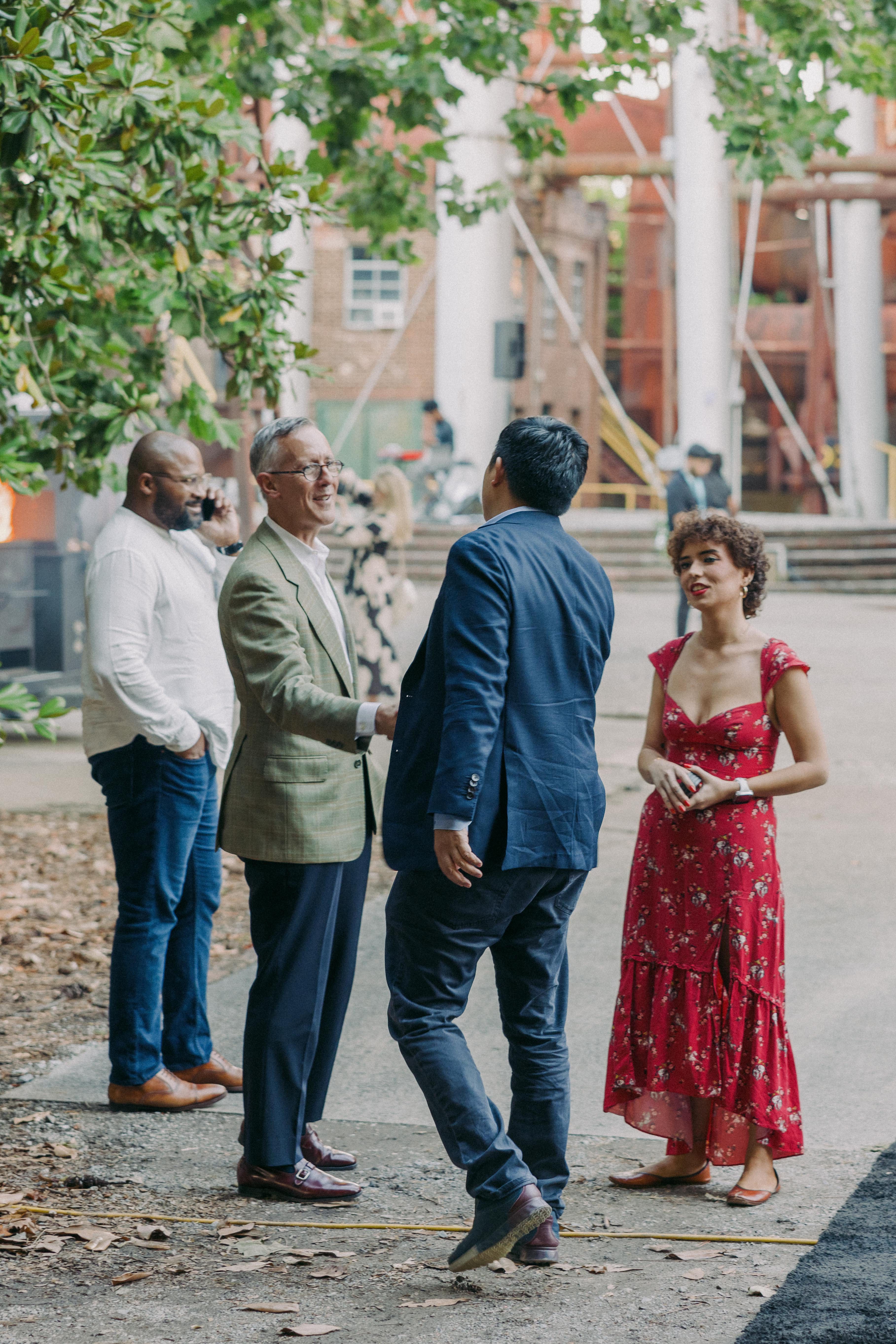 Four people standing outdoors in conversation near large white columns; two men in the foreground are shaking hands, one wearing a green checked blazer and the other a blue suit jacket, while a woman in a red floral dress stands nearby, and another man in a white shirt with jeans is talking on a phone under greenery.