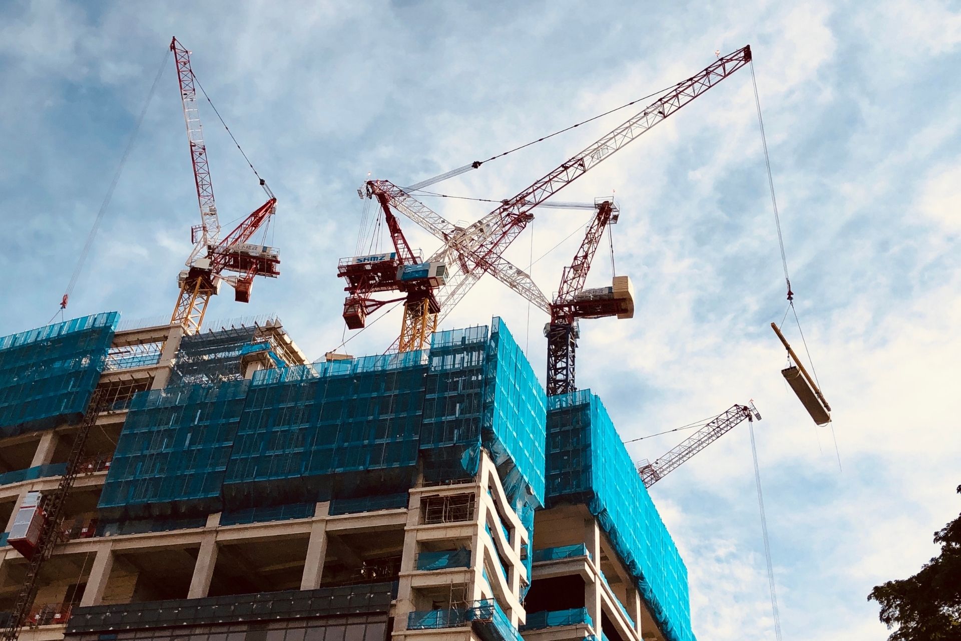 Tower cranes lift materials at a large construction site wrapped in blue mesh, highlighting development under environmental conditions of consent.