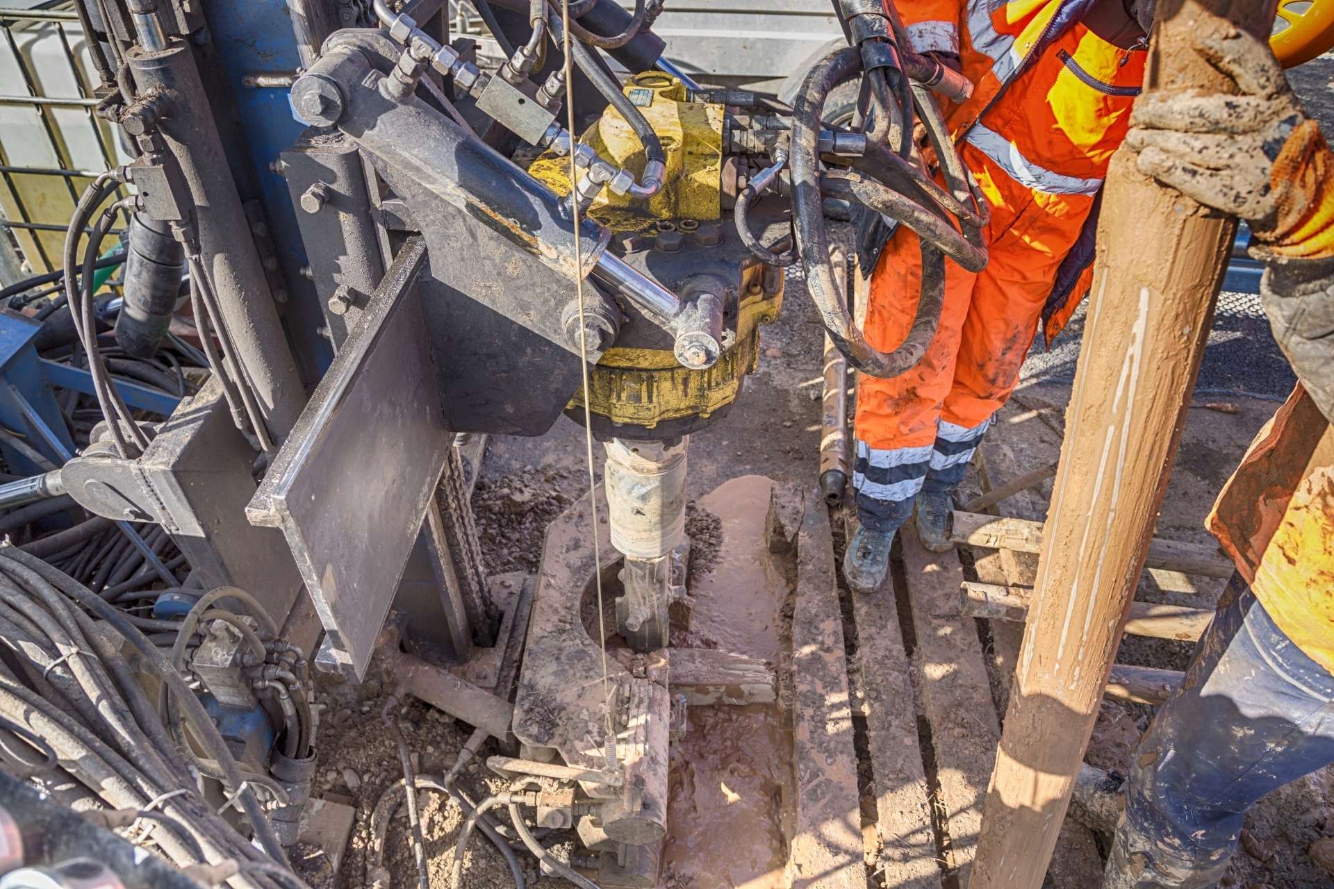 A close-up view of a drilling rig with workers in orange safety gear operating the machinery and handling a large drill bit.