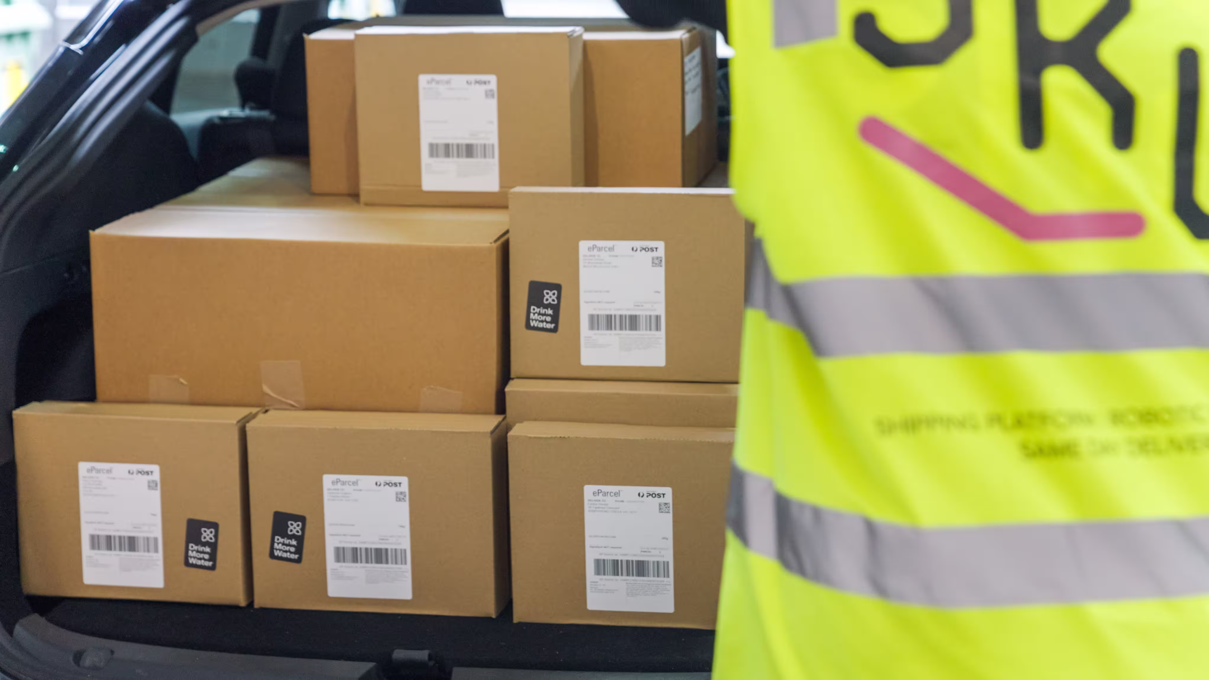 A person wearing a yellow vest is standing next to a stack of brown boxes.