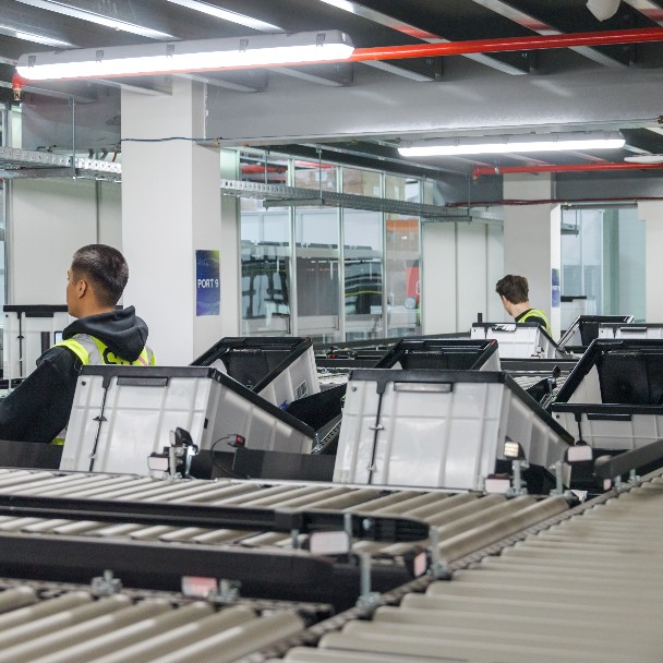 A man in a yellow vest is standing in front of a row of computer monitors.