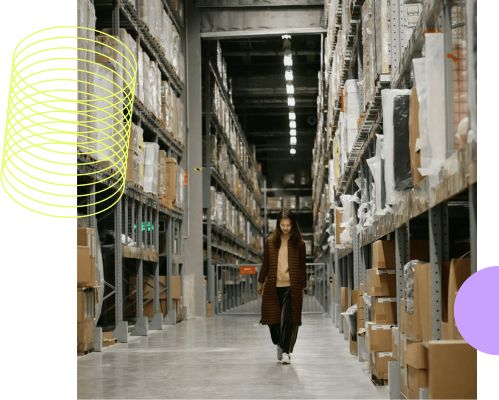 A woman walking down a wide aisle in a large warehouse with shelves stacked with boxes and packages on both sides.