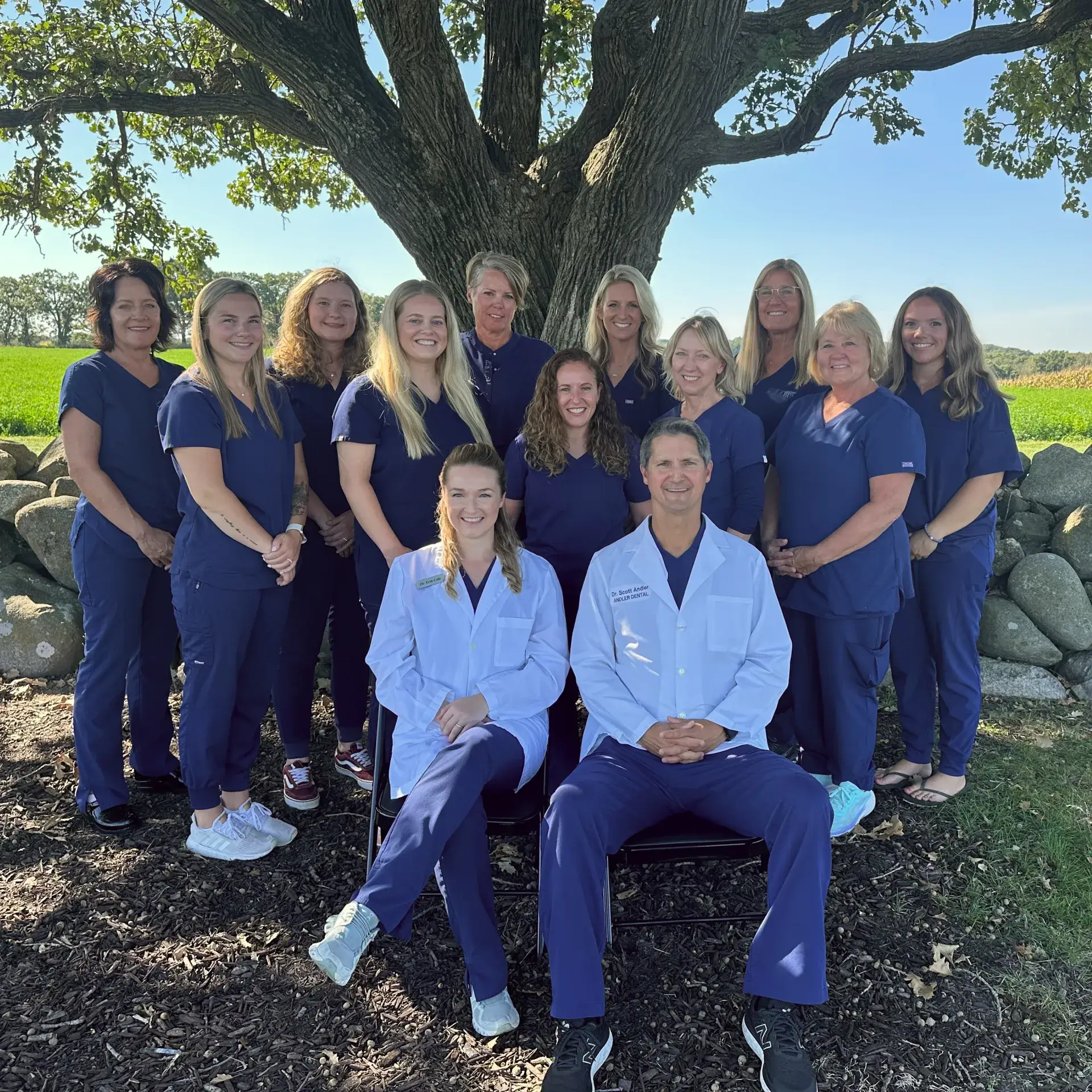 Group of dental professionals in navy scrubs with two seated in white coats, outdoors under a large tree.