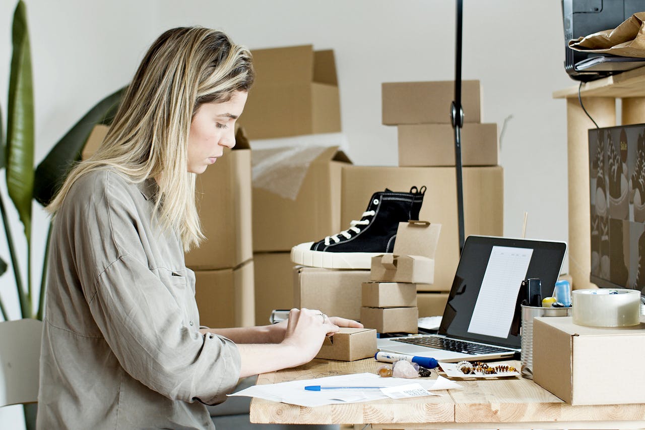 A woman sitting at a desk with a laptop and a stack of brown boxes.