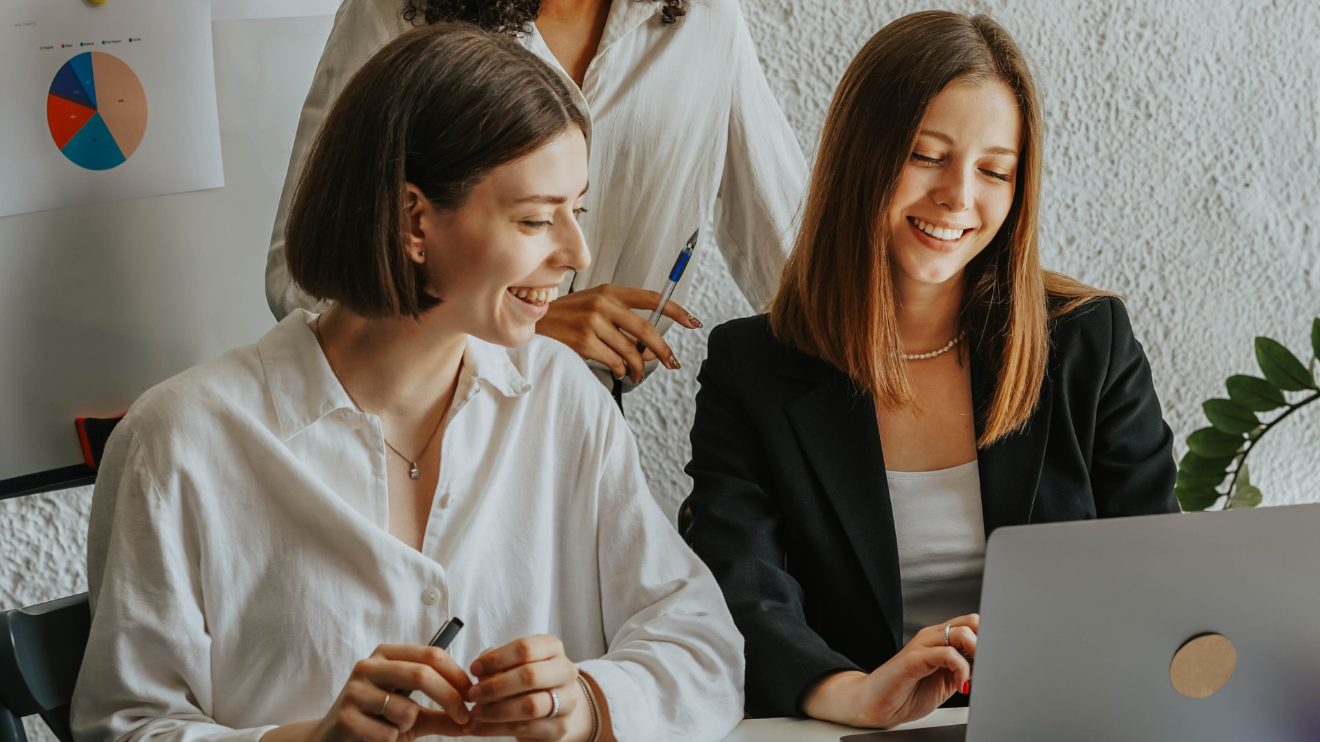 Two women working on a laptop with one of them wearing a black suit.
