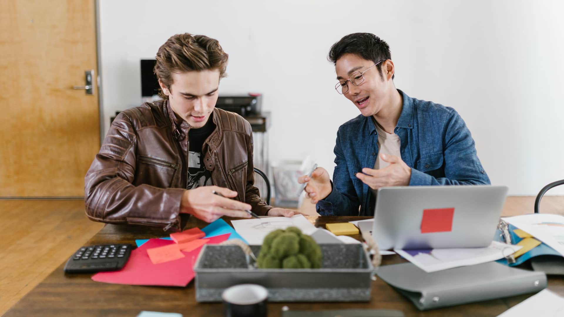 Two men sitting at a table with a laptop and a cup of coffee.