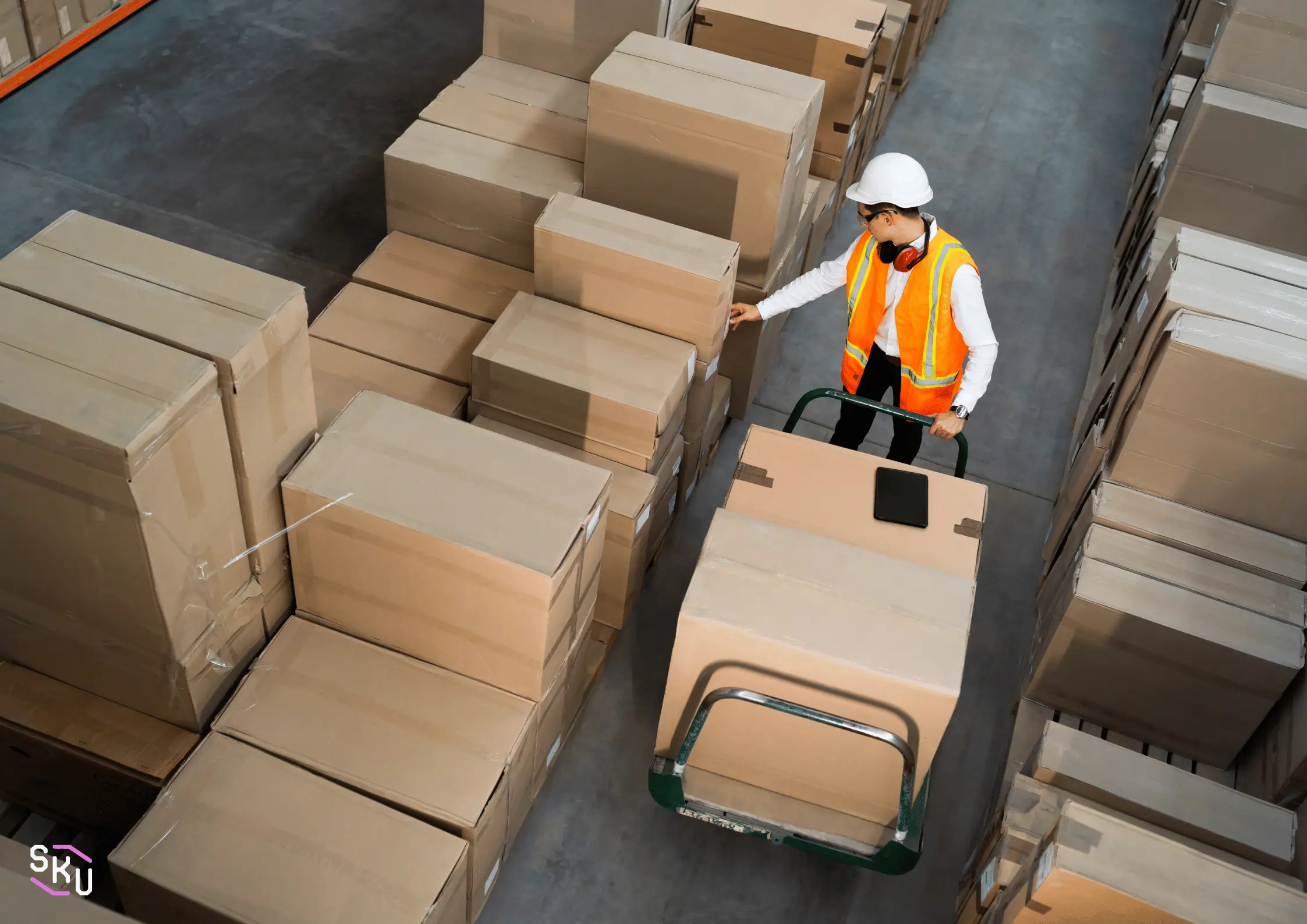 A man in an orange vest is pushing a cart full of brown boxes.
