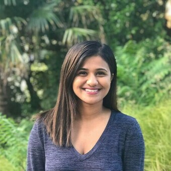 Woman with long dark hair smiling in front of lush green foliage.