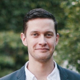 Man in a blazer smiling outdoors with greenery behind him.