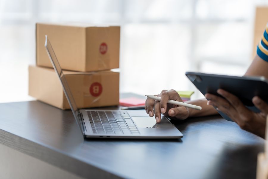 Ecommerce worker using laptop and tablet with parcels nearby, managing online business growth.