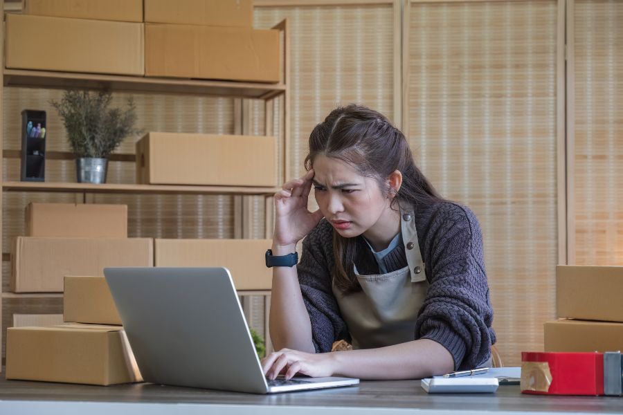 Stressed business owner checking laptop surrounded by boxes, facing stocktake issues.