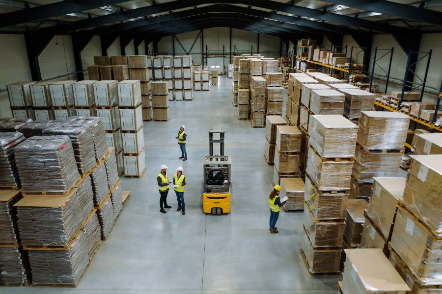 Workers and forklift inside a distribution centre with tall stacks of pallets.