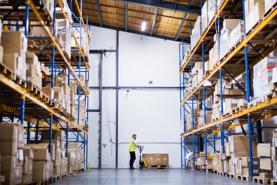 Warehouse worker moving pallet among tall shelves of boxed goods in distribution centre.