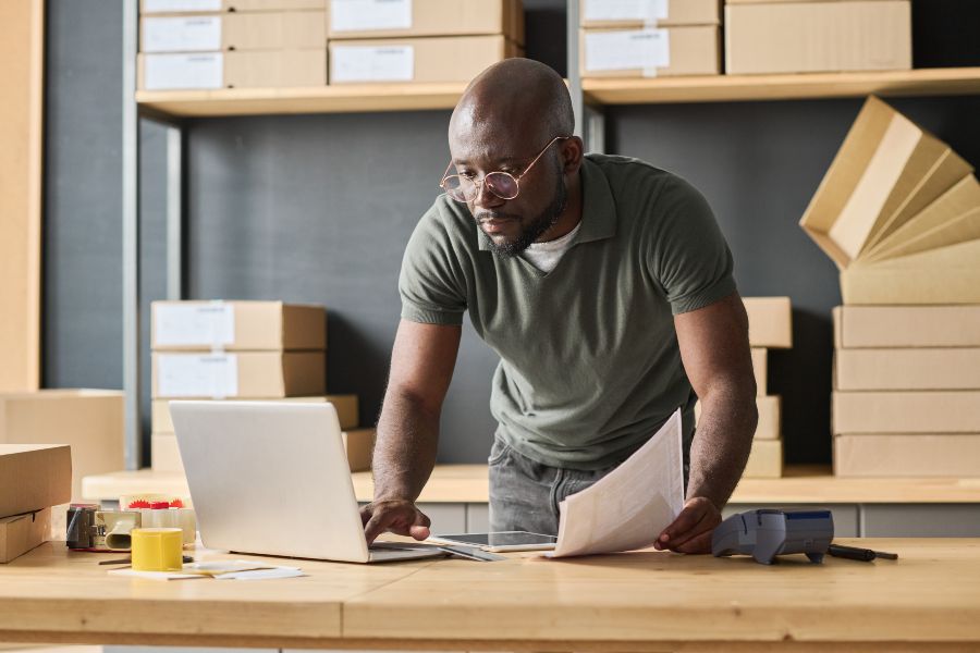 Small business owner stressed while managing orders on a laptop with piles of shipping boxes around.