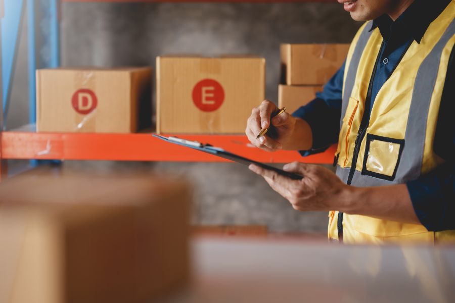 Warehouse worker in safety vest checking inventory with clipboard among labelled boxes.