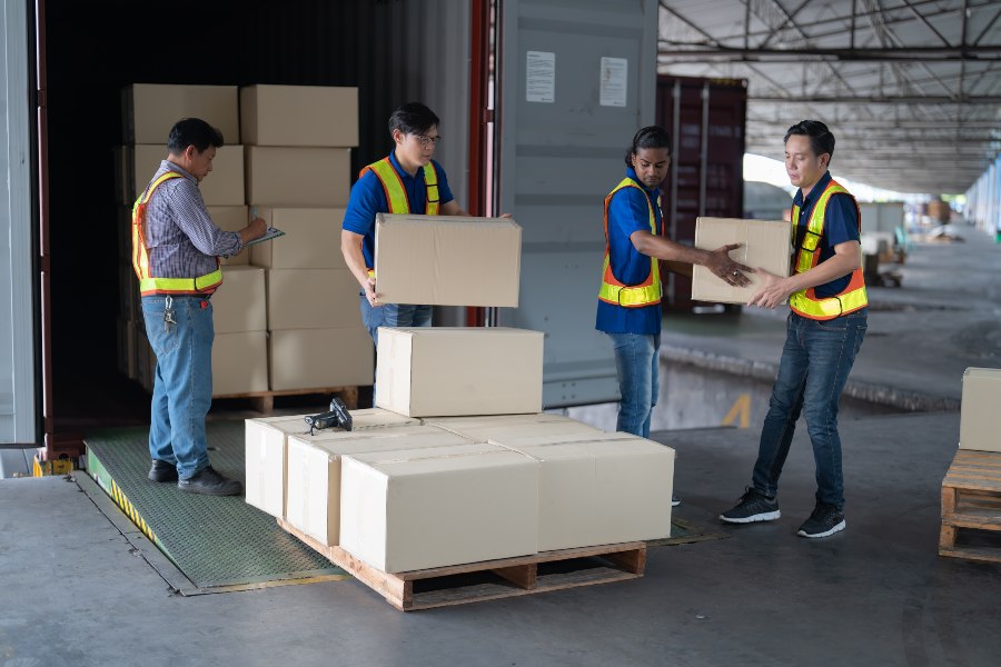 Workers in safety vests loading and checking boxes for international shipping at a warehouse dock.