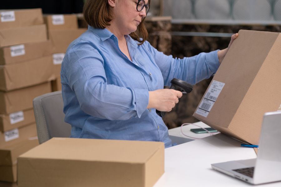 Woman scanning a shipping box with a barcode scanner in a packaging workspace.