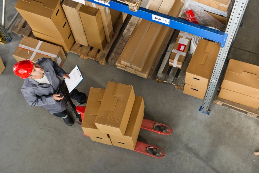 Worker with a red hard hat checking boxes on a pallet jack in a warehouse.