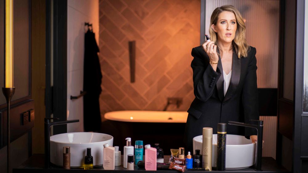A woman in a black suit is applying makeup in front of a table full of cosmetics.