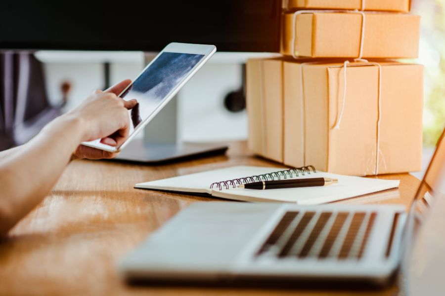 Person using a tablet to manage orders with shipping boxes and notebooks on a desk