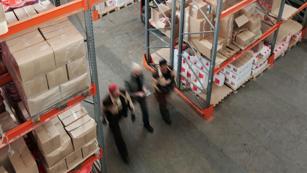 A man in a brown jacket is walking in front of a shelf of boxes.