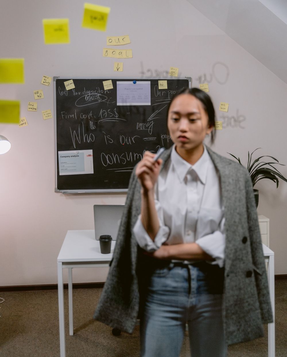 A woman in a white shirt and gray jacket is standing in front of a table with a laptop and a bunch of yellow sticky notes.